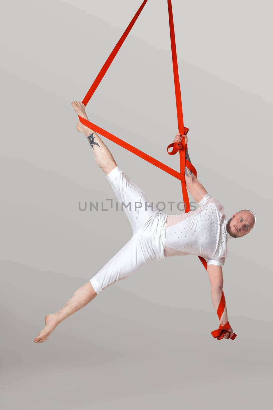 Sportsman performance on a red canvases. Bearded male in a light sport suit is doing an acrobatic elements hanging on a rope in a studio isolated on white background. Dancing in the air with balance.