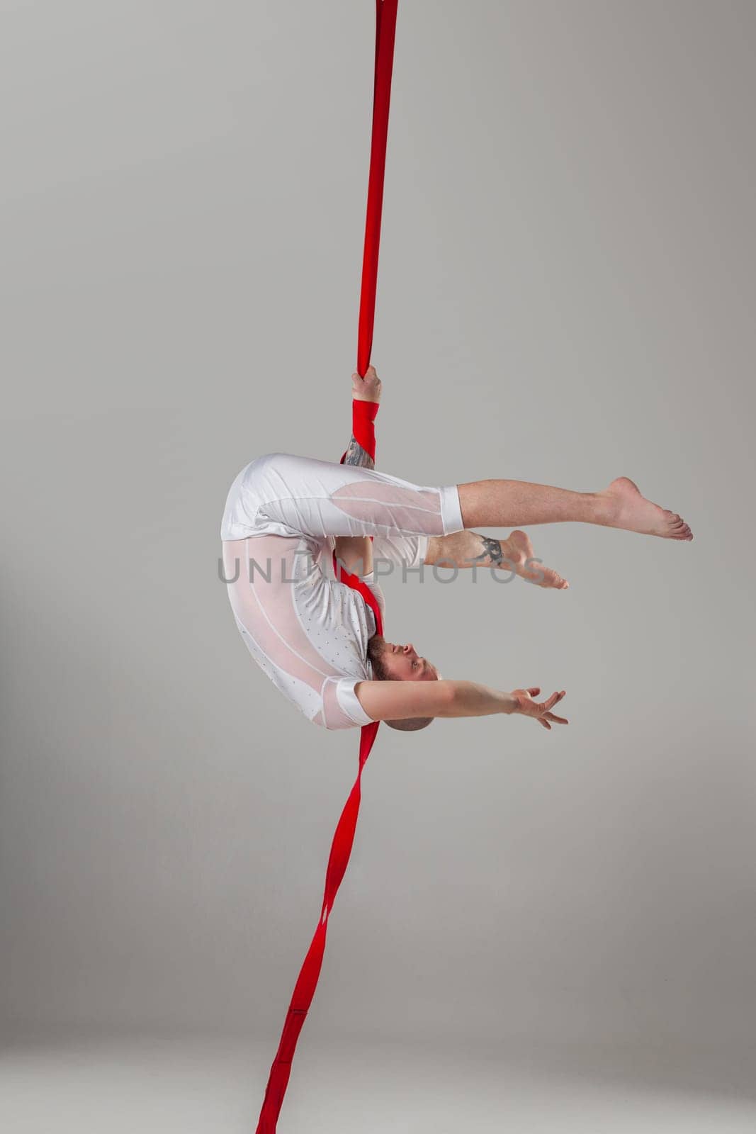 Gymnast performance on a red canvases. Athletic fellow in a light sport suit is doing an acrobatic elements hanging upside down on a rope in a studio isolated on white background. Dancing in the air with balance.