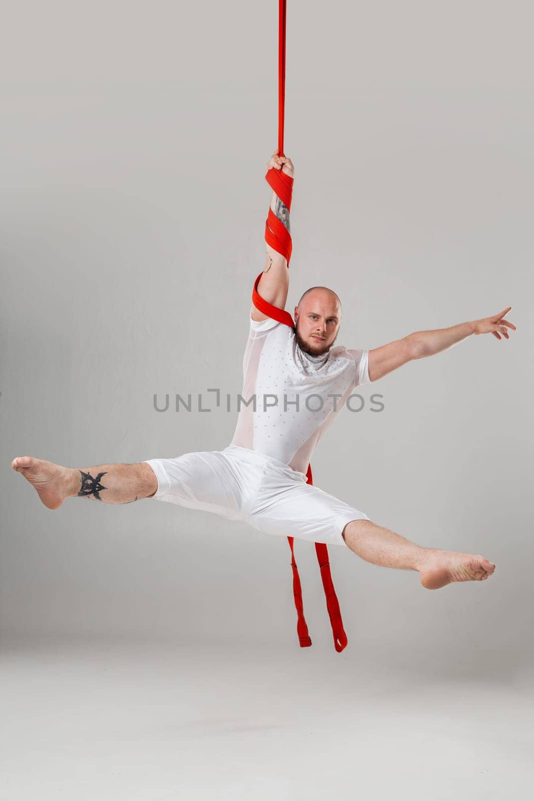 Gymnast performance on a red canvases. Athletic man in a light sport suit is doing an acrobatic elements hanging on a rope in a studio isolated on white background. Dancing in the air with balance.