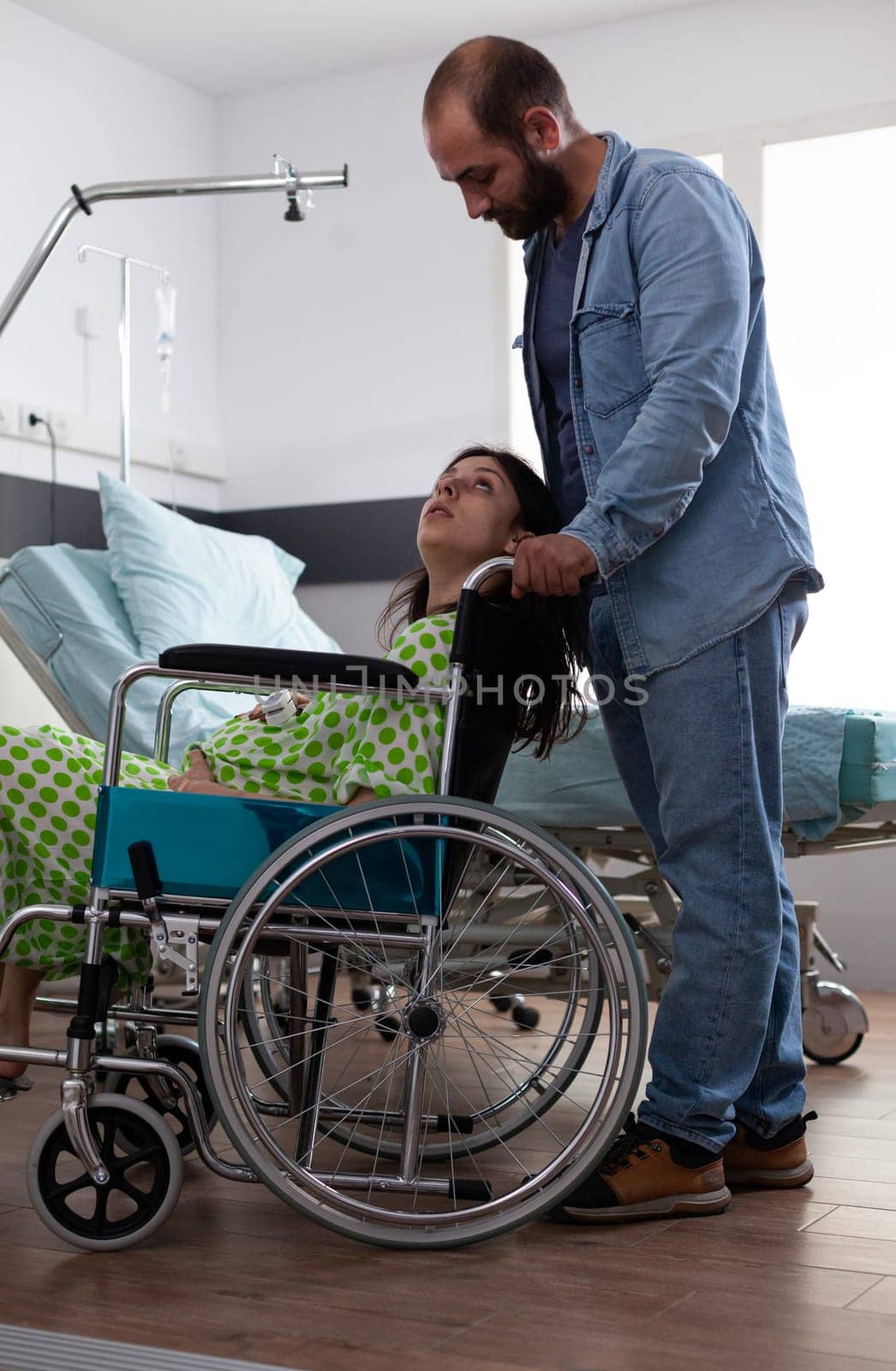 Husband taking pregnant woman in wheelchair to maternity room, preparing for baby delivery in hospital ward. Young couple getting ready for childbirth, discussing parenthood. Birth concept