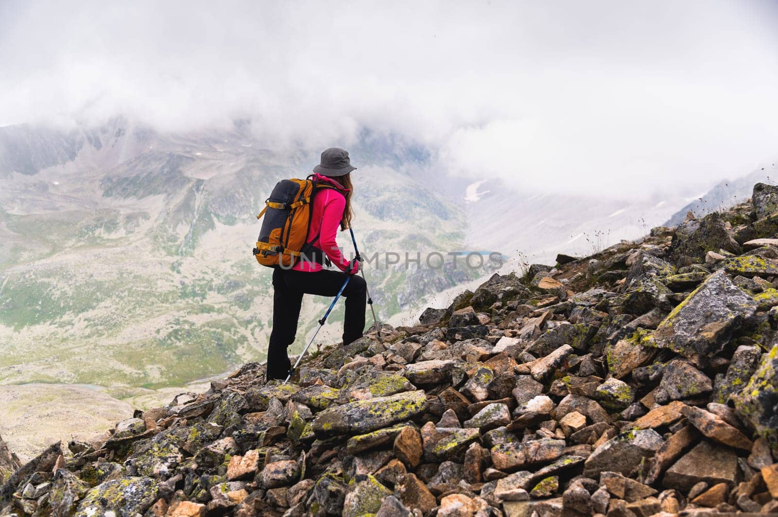 A woman traveler goes to the top of the mountain. A girl with trekking poles and a backpack stopped to admire the fog approaching her in the mountains.