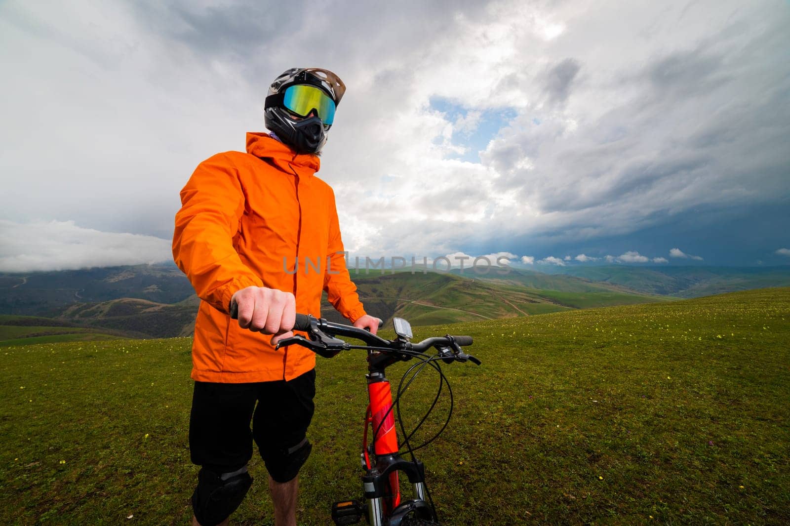 a man in a helmet and protection stands with a bicycle. on a green hill among the mountains during a break between riding off the road.