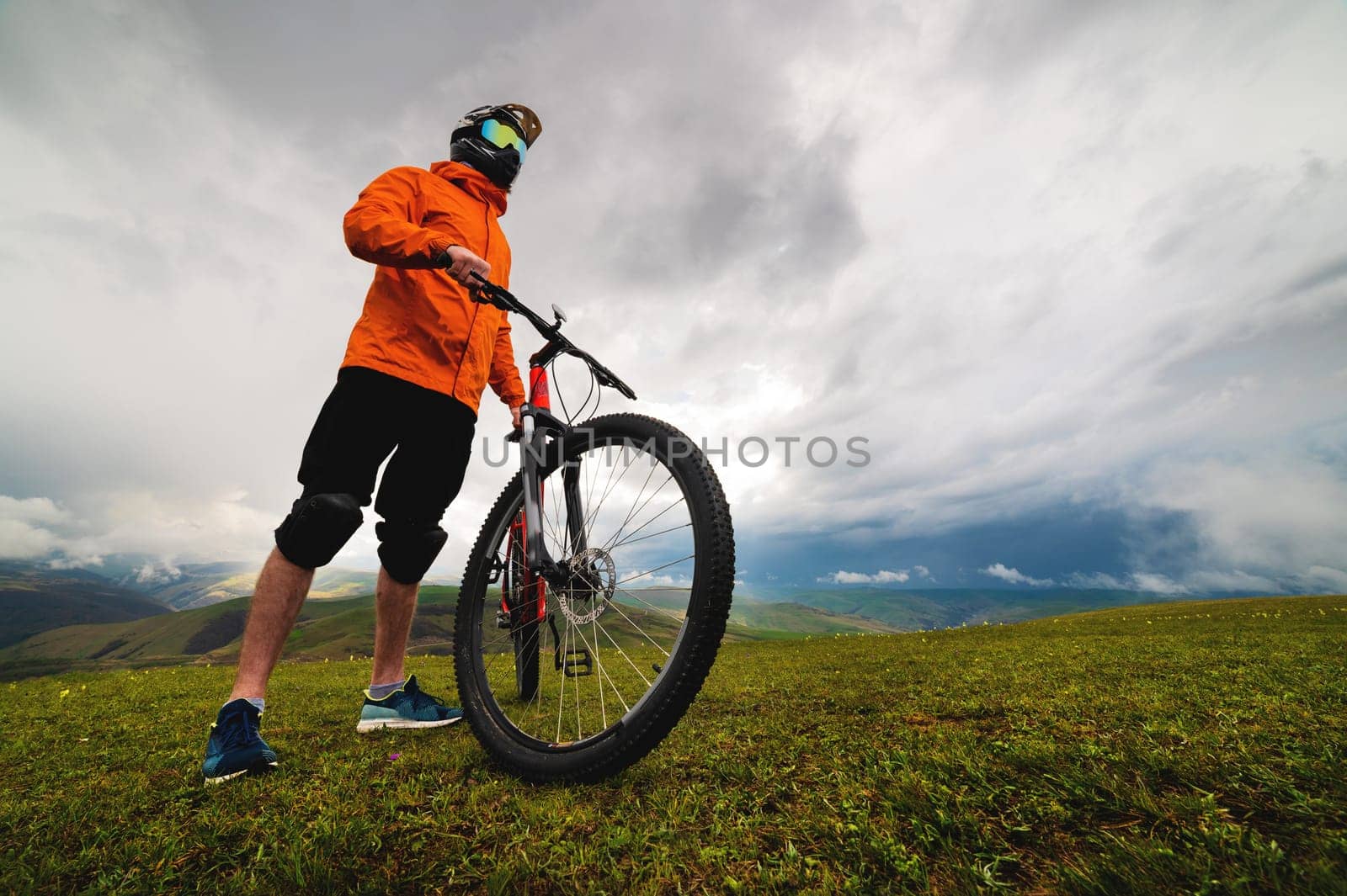 man in a helmet and protection stands with a bicycle on a green hill among the mountains during a break between riding off the road by yanik88