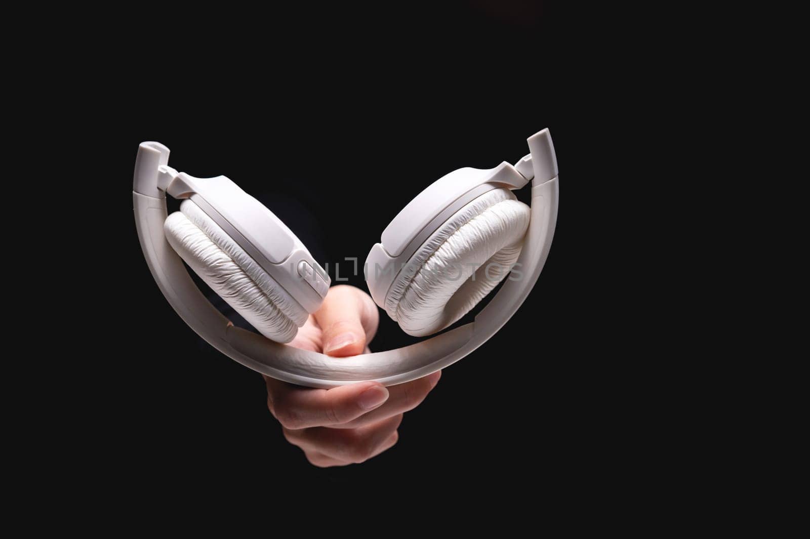 A hand holds headphones on a black background. Modern white headphones