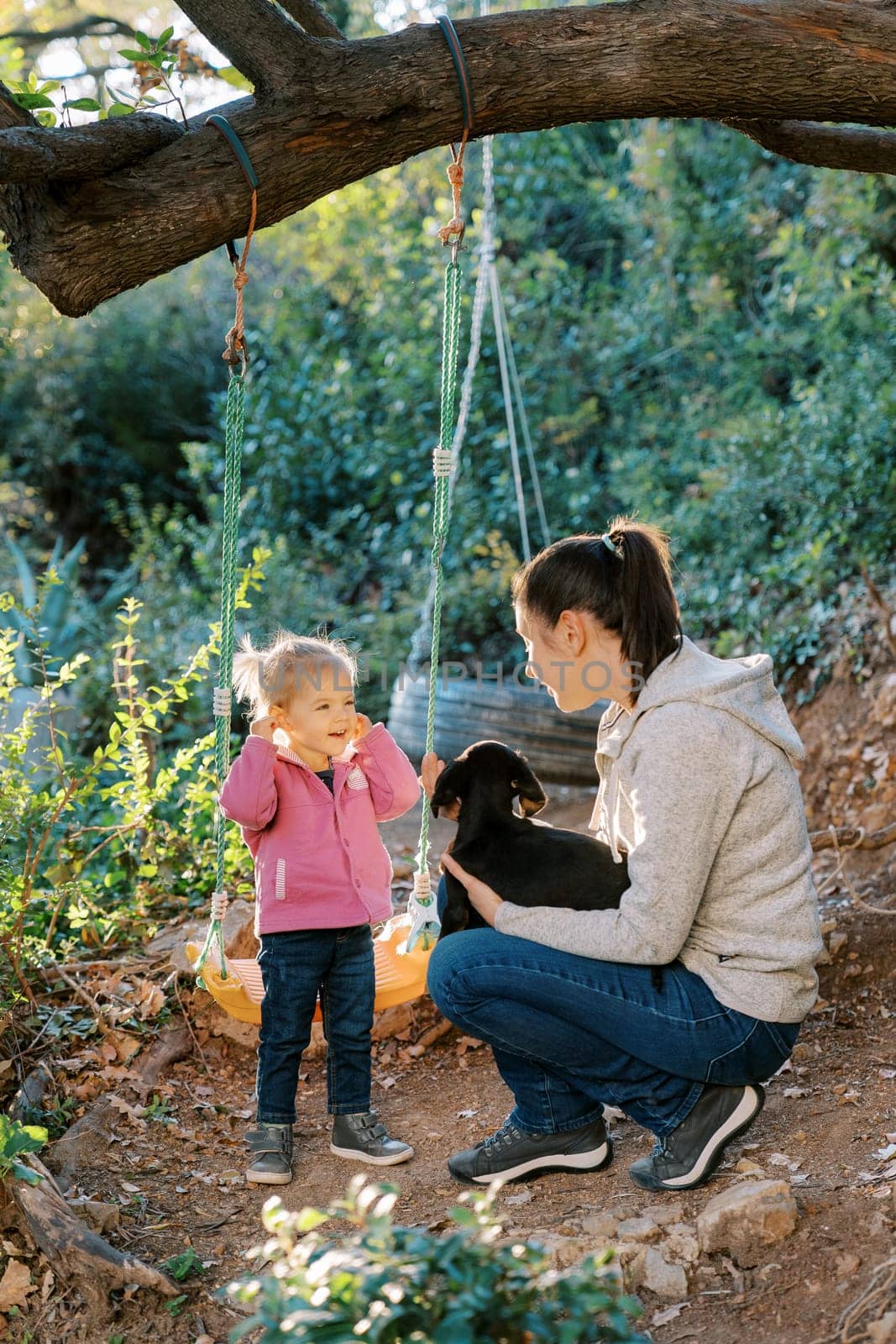 Little smiling girl stands near a swing and watches her mom petting a puppy on her lap. High quality photo