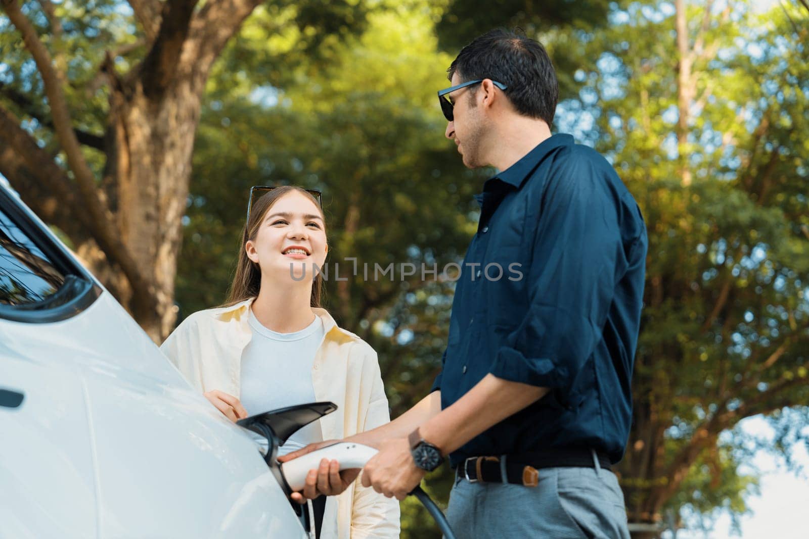 Young couple recharge electric car's battery from charging station in outdoor green city park in springtime. Rechargeable EV car for sustainable environmental friendly urban travel lifestyle.Expedient