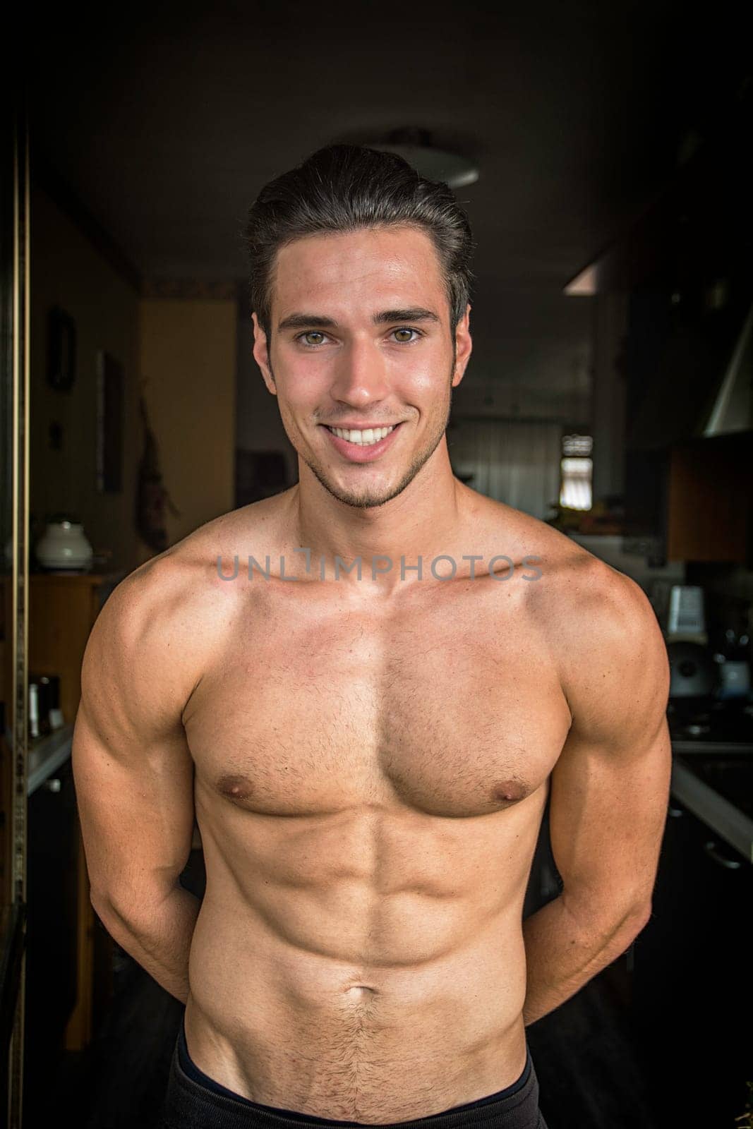 A young handsome athletic muscular man with no shirt posing for a picture inside a house, looking at camera smiling