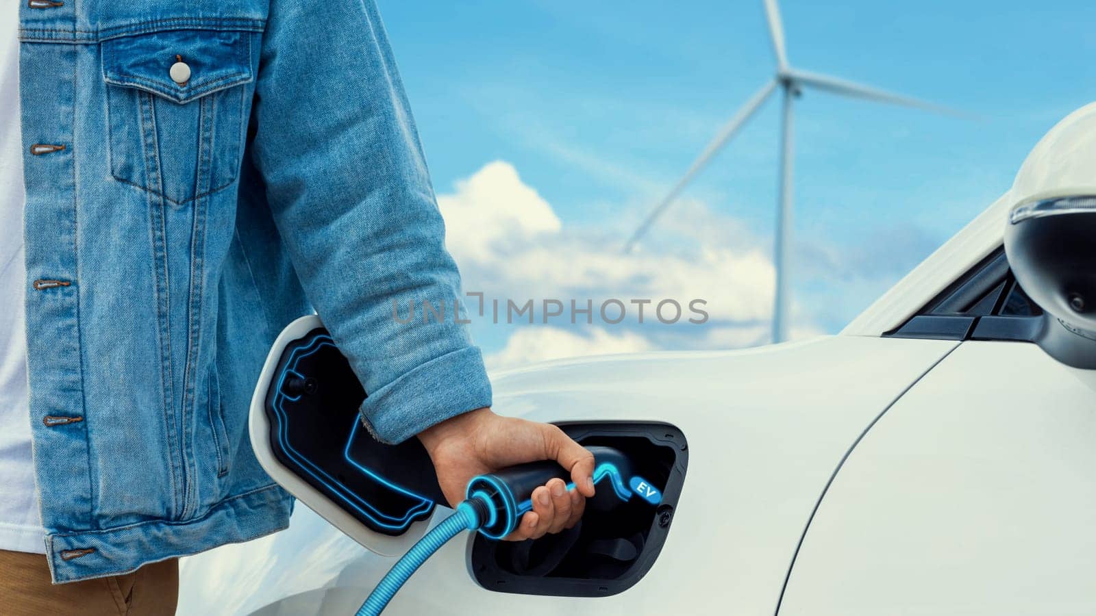Hand insert EV charger and recharge electric car from charging station with wind turbine generator background. Smart eco-friendly EV car. Technological advancement of alternative energy. Peruse