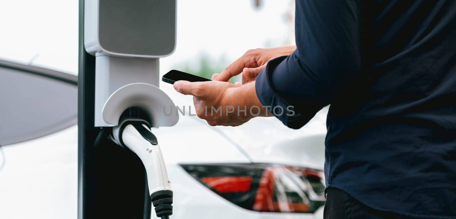 Man using smartphone online banking application to pay for electric car battery charging from EV charging station during vacation road trip at national park or summer forest. Panorama Exalt