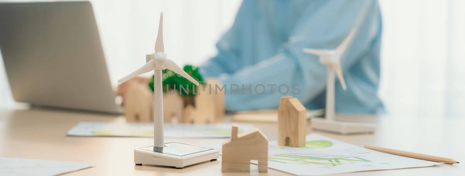 Windmill model represented using clean energy and wooden block represented eco house was scatter around on the table in front of businessman working on laptop. Blurring background. Delineation.