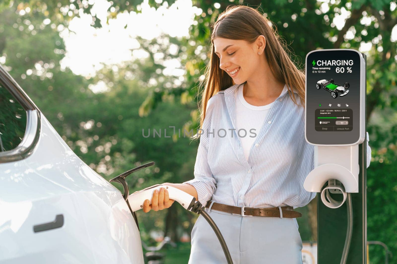 Young woman recharging battery for electric car during road trip travel EV car in natural forest or national park with sunrise lighting. Eco friendly travel during vacation and holiday. Exalt