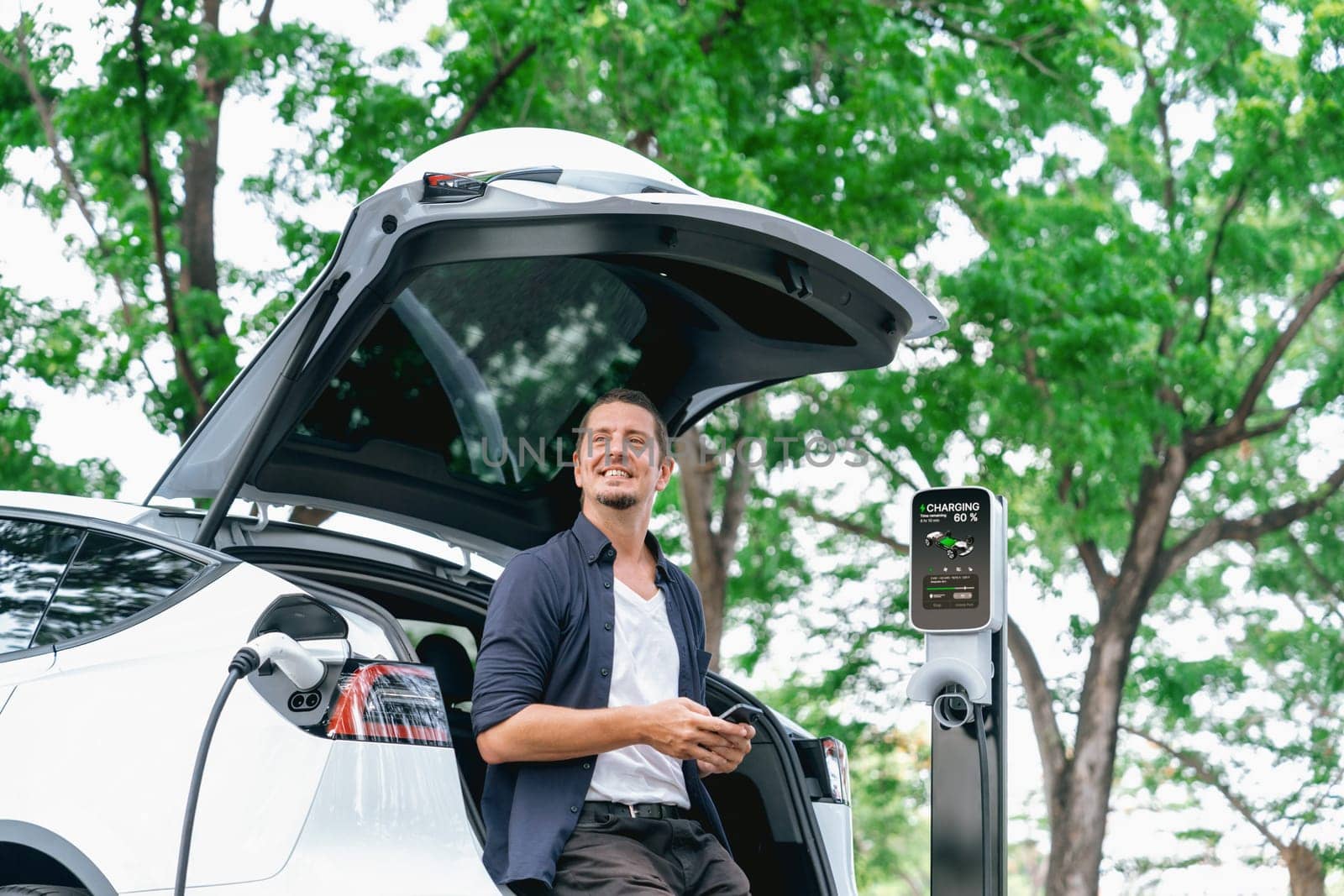 Man using smartphone online banking application to pay for electric car battery charging from EV charging station during vacation holiday road trip at national park or summer forest. Exalt