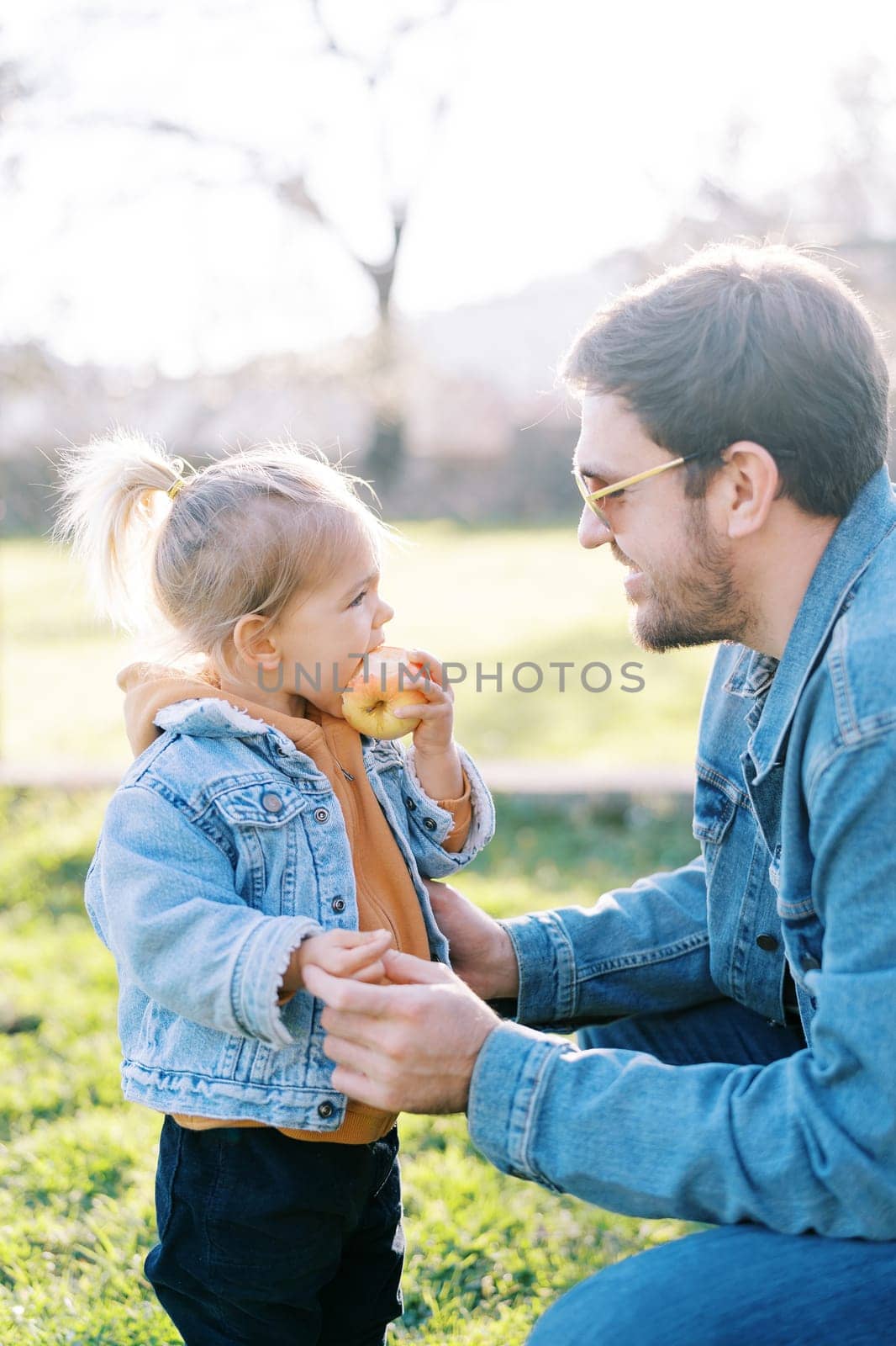 Smiling dad squatted opposite to a little girl eating an apple on a green meadow by Nadtochiy