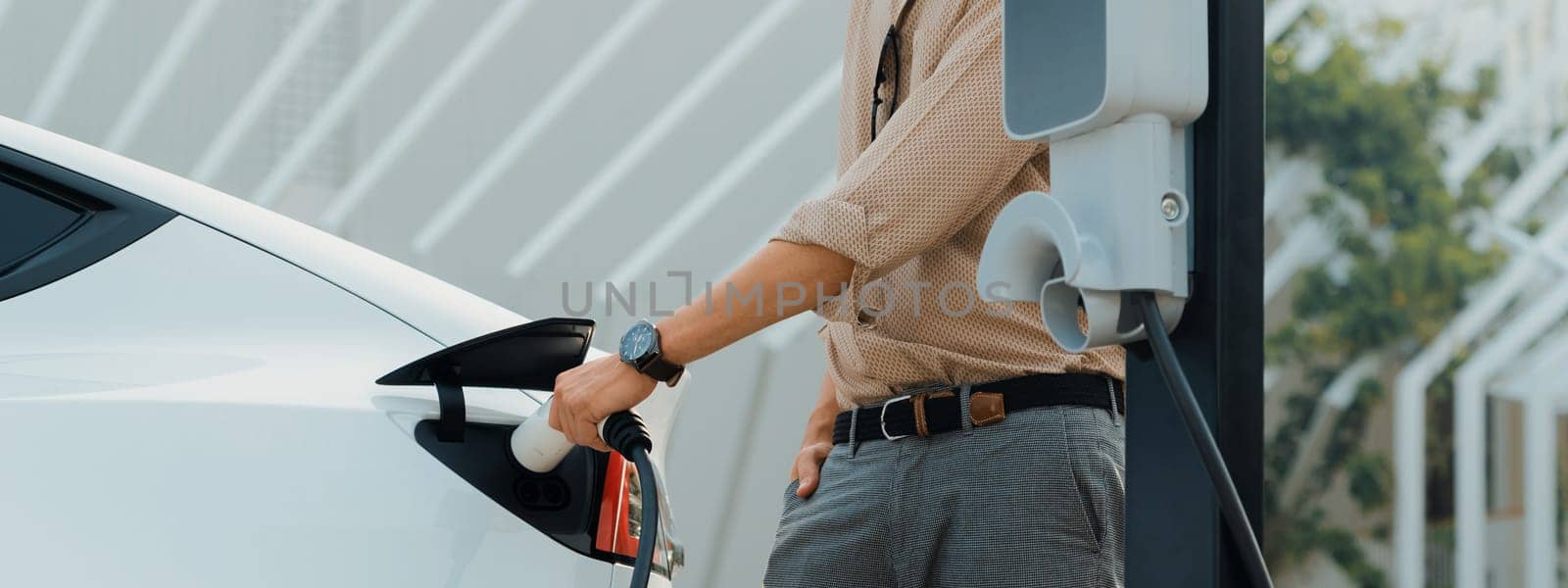 Young man recharge electric car's battery from charging station in city commercial parking lot. Rechargeable EV car for sustainable environmental friendly urban travel. Panorama Expedient