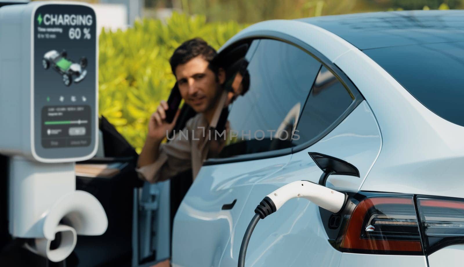 Young man recharge EV electric vehicle at green city mall parking lot while talking on phone. Sustainable urban lifestyle for eco friendly EV car with battery charging station. Panorama Expedient