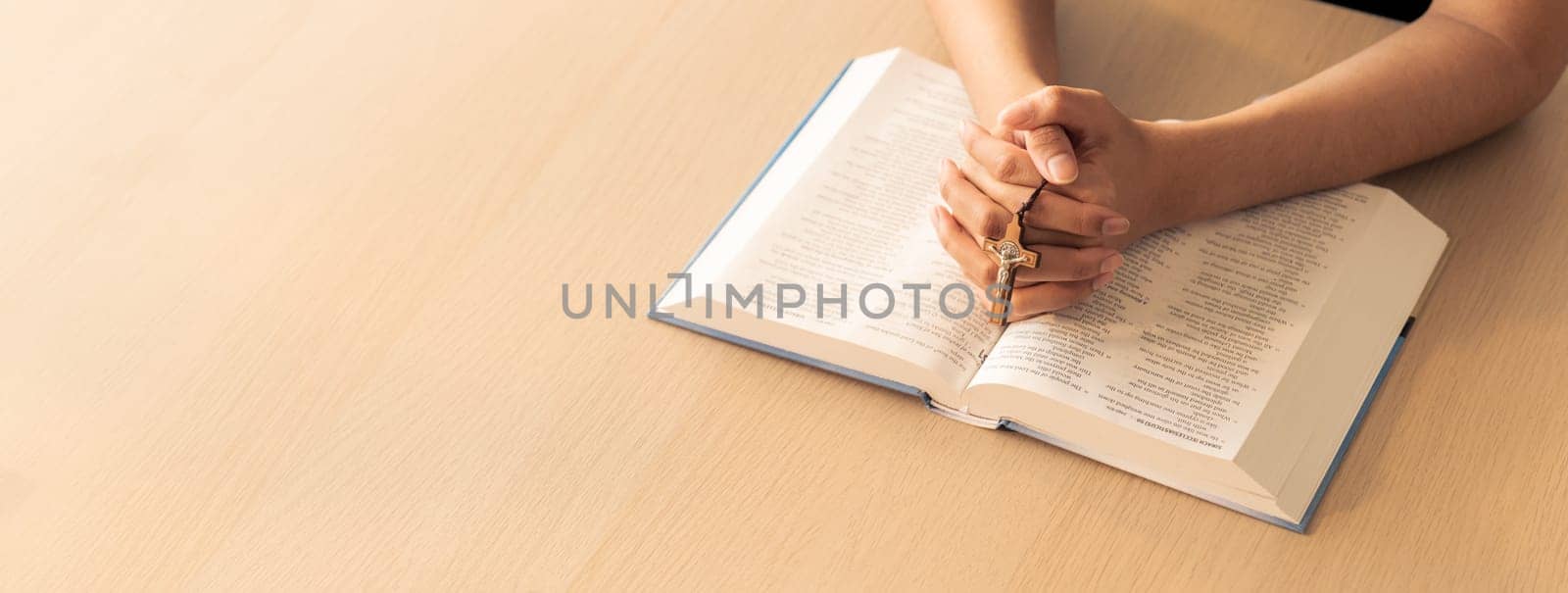 Cropped image of praying male hand holding cross on holy bible book at wooden table. Top view. Concept of hope, religion, faith, christianity and god blessing. Warm and brown background Burgeoning.