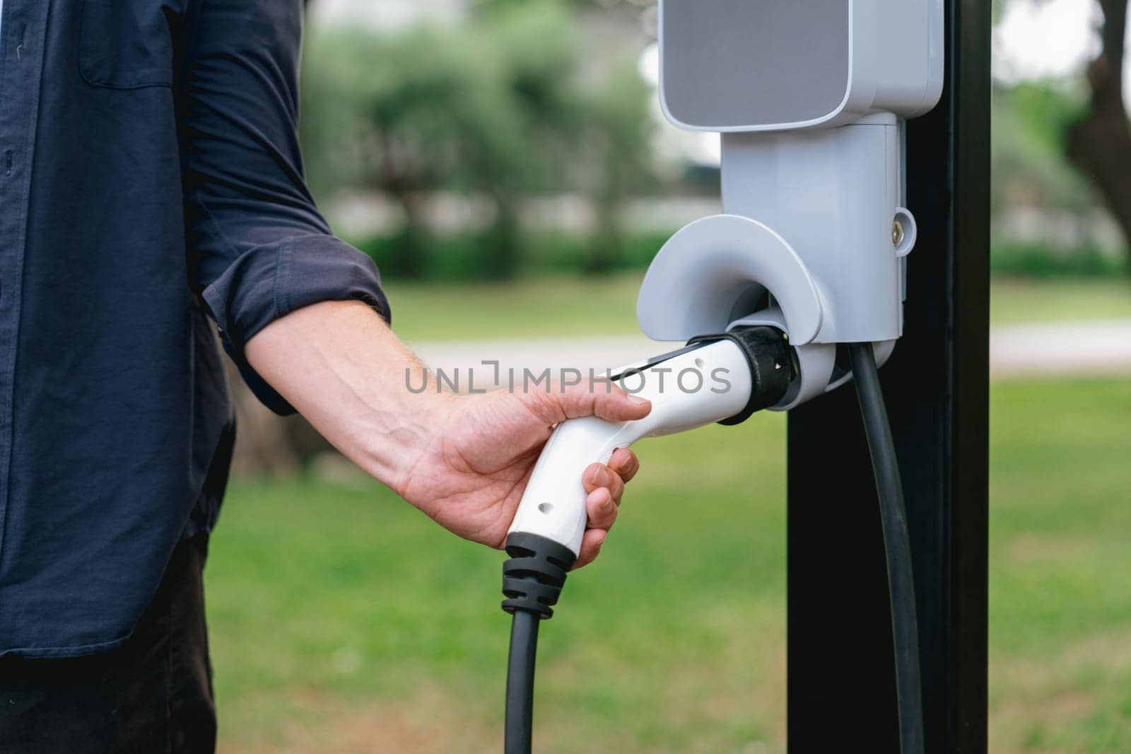 Man recharge EV electric vehicle's battery from EV charging station in outdoor green city park scenic. Eco friendly urban transport and commute with eco friendly EV car travel. Exalt