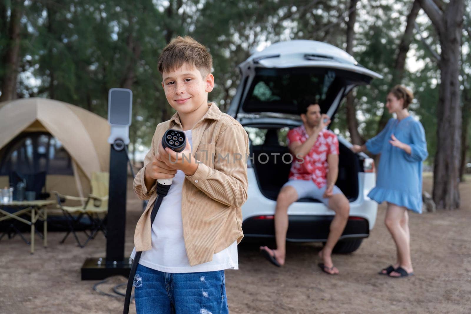 Little boy holding EV charger and point at camera with his family sitting on the trunk in background. Road trip travel with alternative energy charging station for eco-friendly car concept. Perpetual