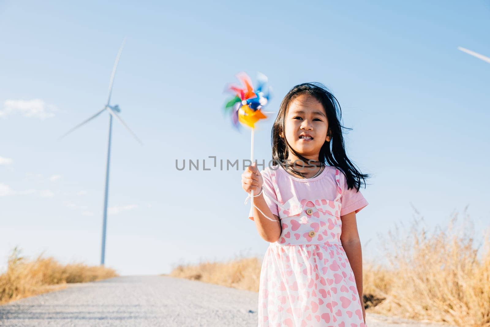 A joyful little girl smiling and running with pinwheels near windmills. Embracing clean energy through playful education in a picturesque wind turbine setting under a beautiful sky.