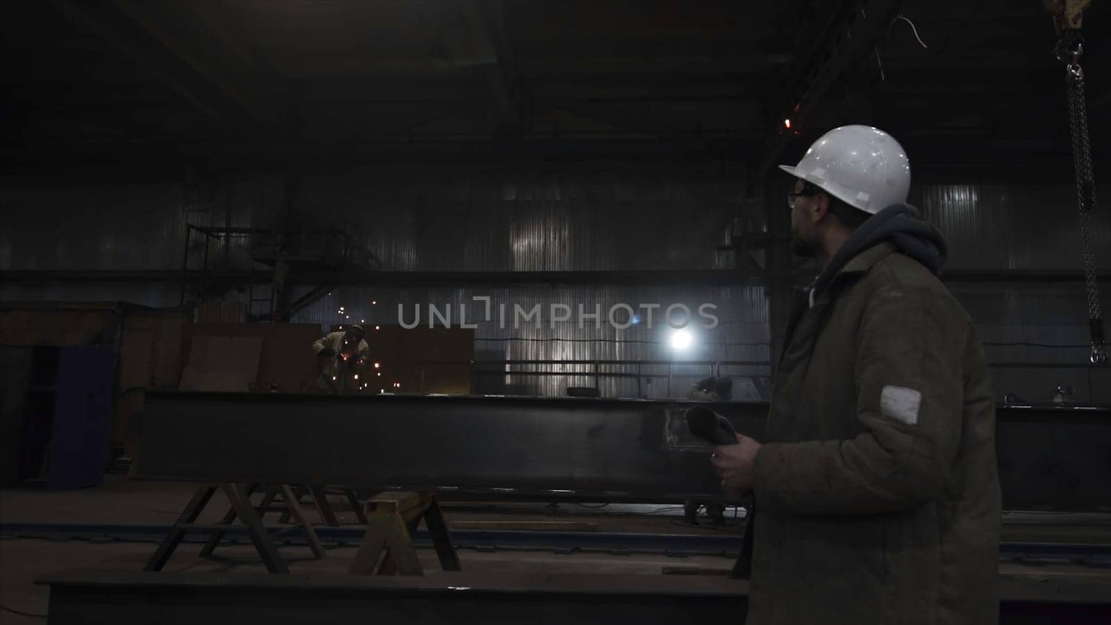 Man walks in helmet at factory on background of workers. Clip. Man looks at work of factory workers. Man walks on background of worker with sparkling welding.