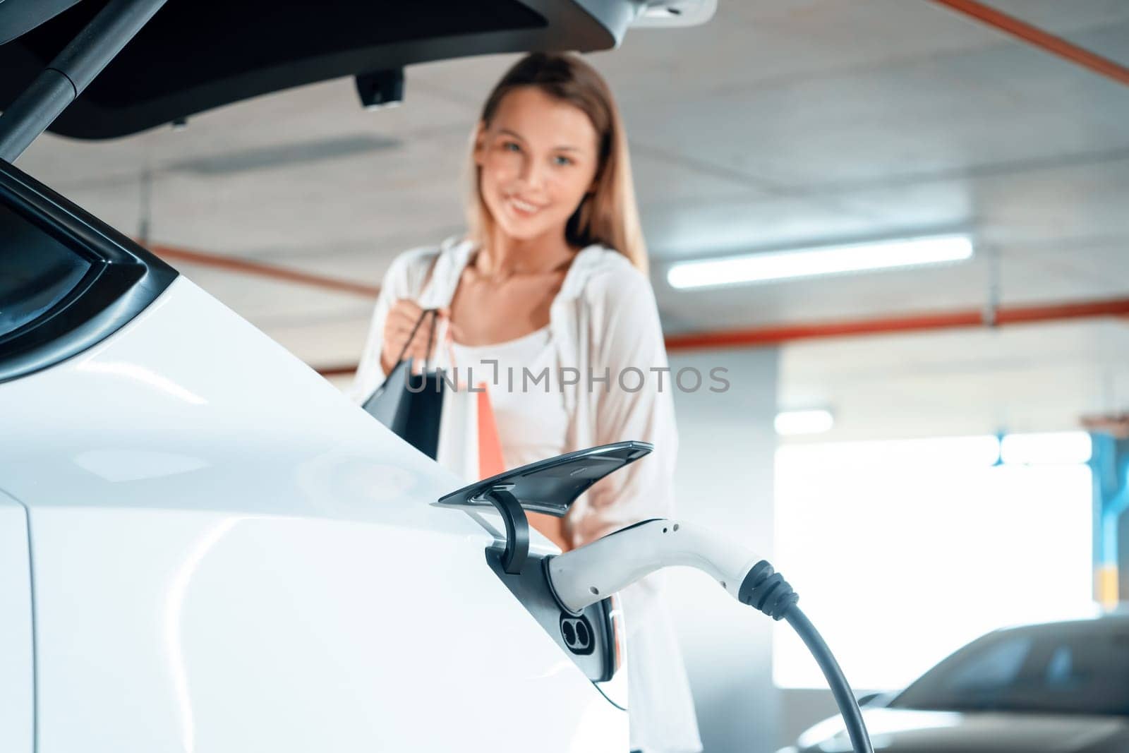 Young woman travel with EV electric car to shopping center parking lot charging in downtown city showing urban sustainability lifestyle by green clean rechargeable energy of electric vehicle innards