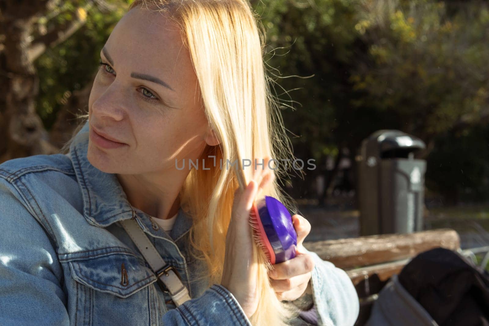 beautiful blonde young adult woman combing her hair while sitting on a bench in the park by PopOff
