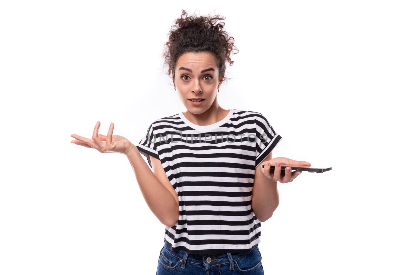 young charming curly brunette woman in a striped t-shirt shrugs her shoulders holding a phone.
