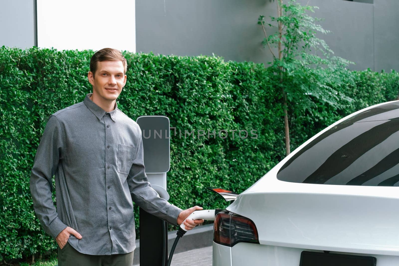 Young man travel with EV electric car charging in green sustainable city outdoor garden in summer shows urban sustainability lifestyle by green clean rechargeable energy of electric vehicle innards