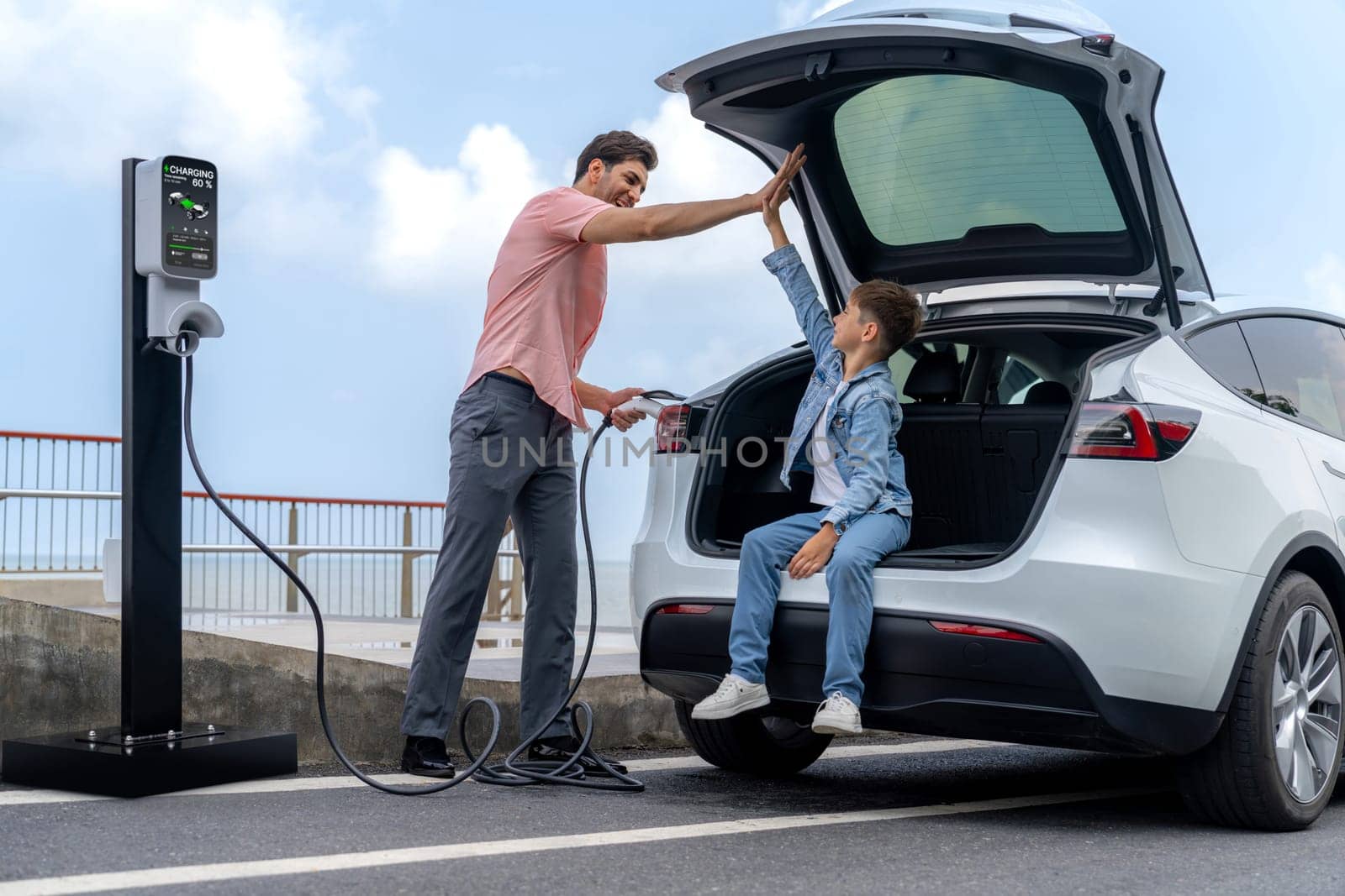 Family road trip vacation traveling by the sea with electric car, father and son high five after reach destination at EV charging station by the seashore. Eco-friendly car for environment. Perpetual