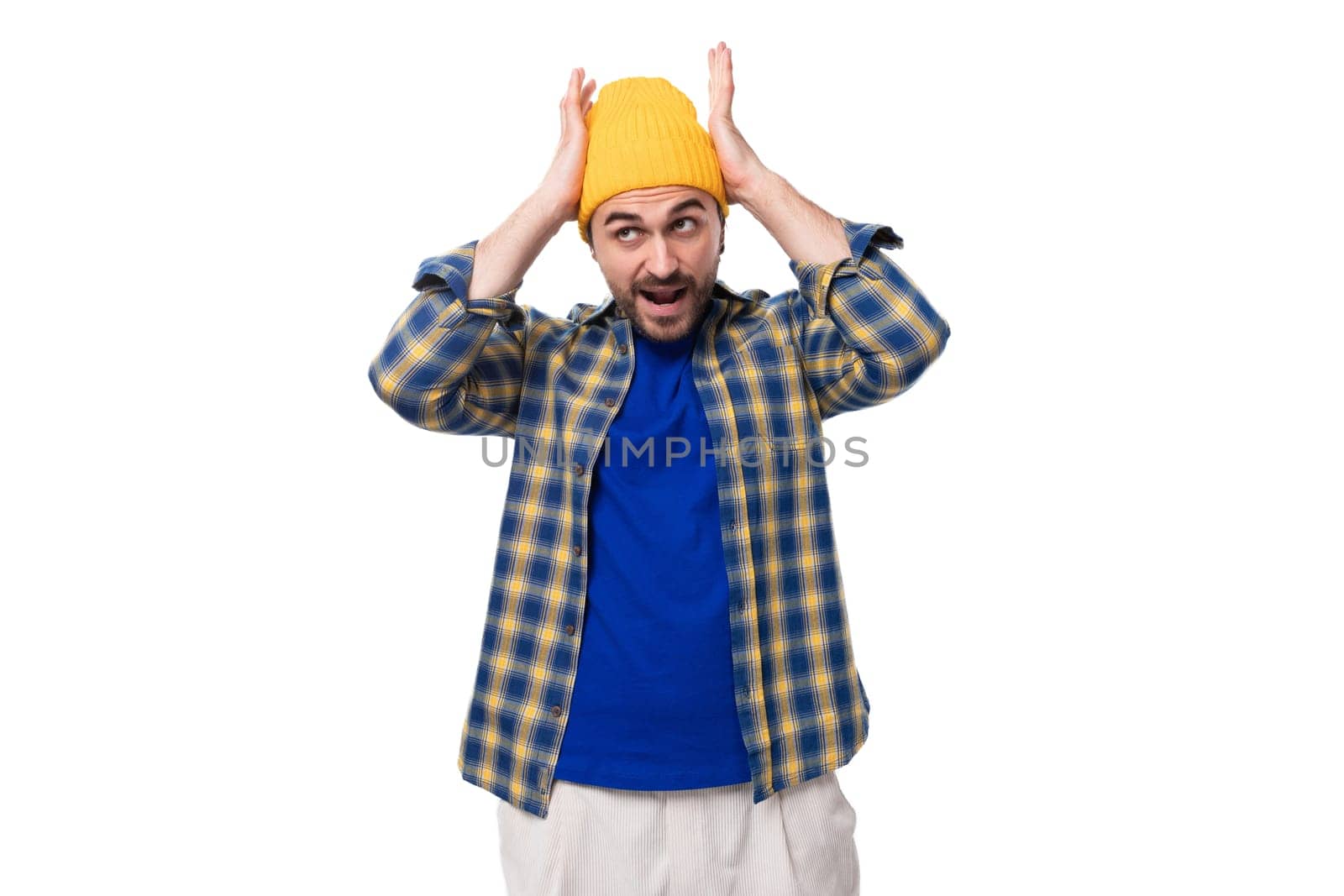 young fashionable brunette man with a brutal beard and mustache in a hat and shirt holding his head on a white background with copy space by TRMK