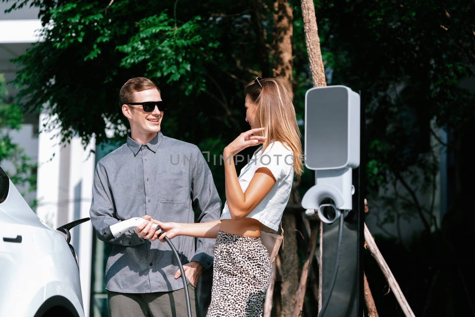 Young couple travel with EV electric car charging in green sustainable city outdoor garden in summer shows urban sustainability lifestyle by green clean rechargeable energy of electric vehicle innards