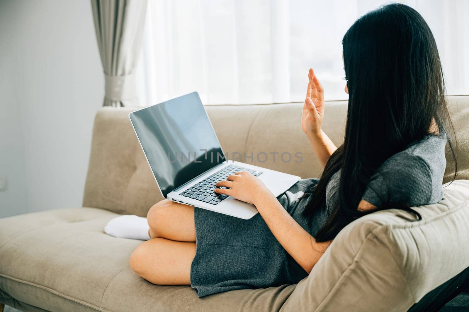 Happy woman on sofa uses laptop for video conferencing waving hi. Engaged in distant meeting discussing and smiling. Modern online communication and connection.