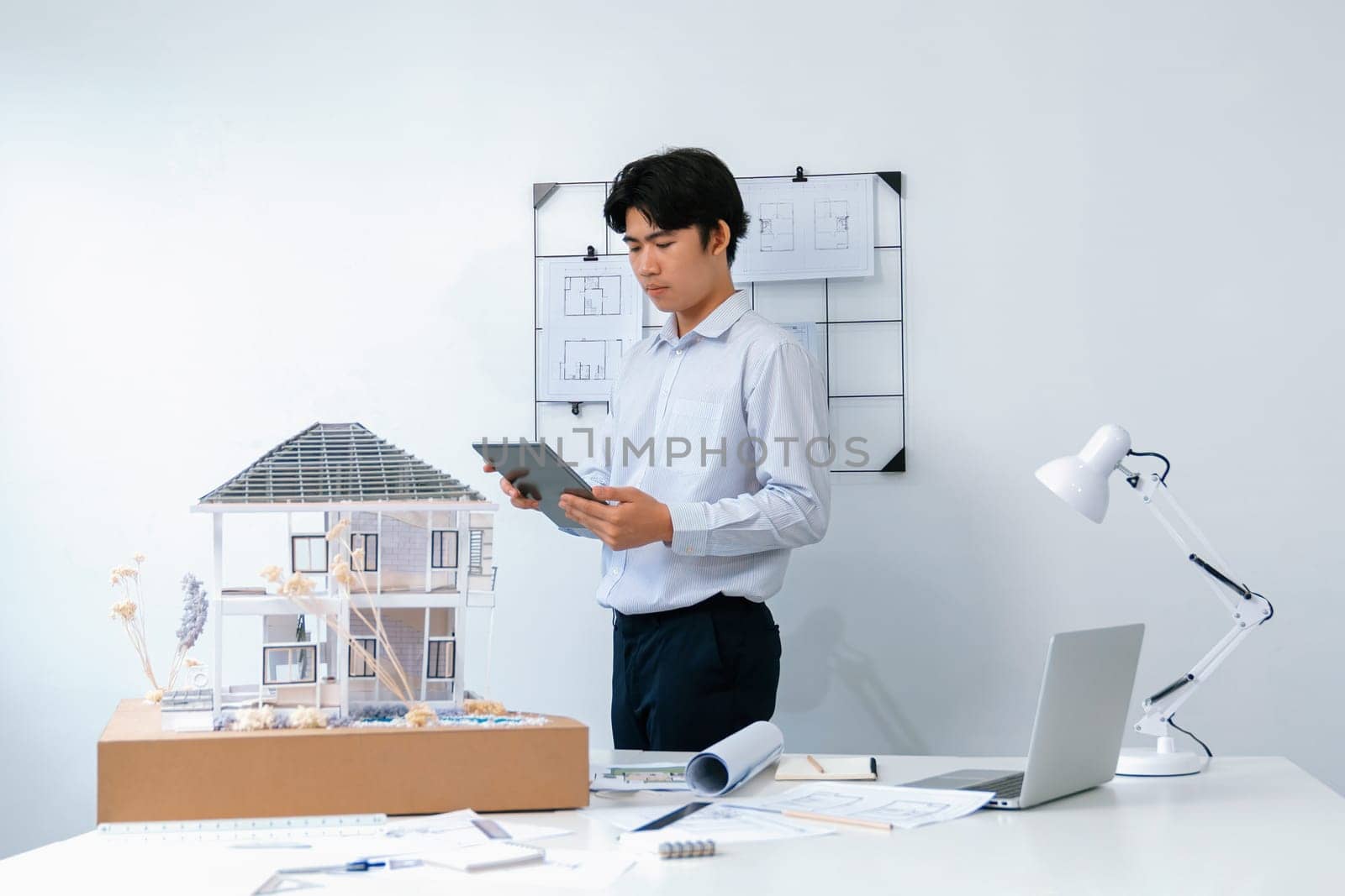 Professional handsome asian project manager using tablet to check house model while inspect house model structure carefully at office with blueprint hanged behind. Creative design. Immaculate.