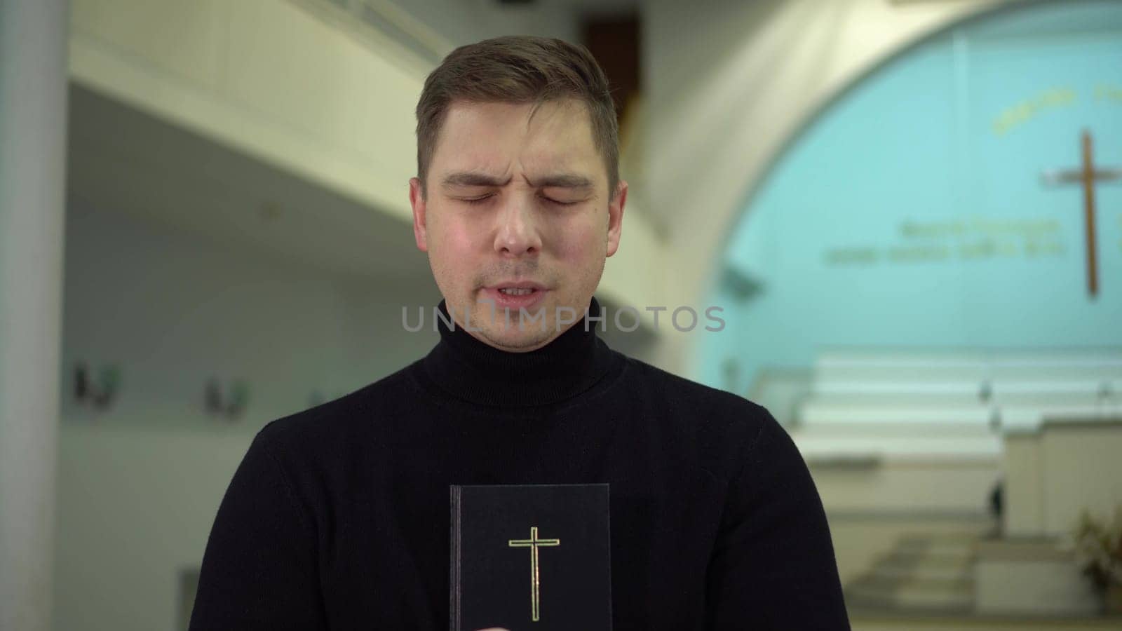 A young man prays holding a bible in his hands in a church against the background of a cross. A Protestant man stands in a church and prays. by Puzankov