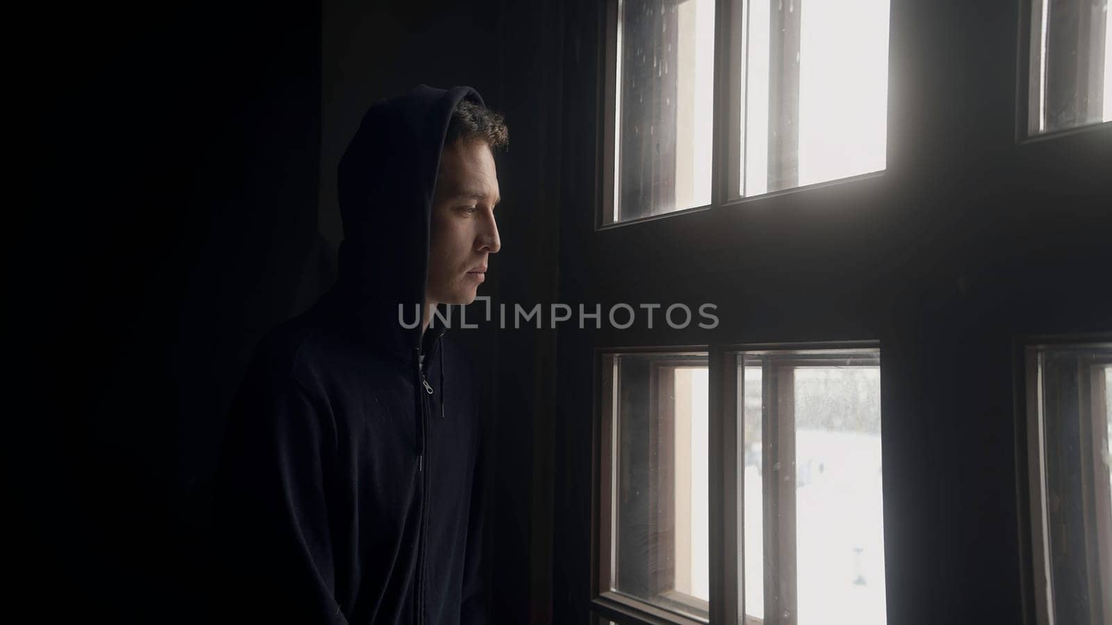 A thoughtful man at the window. Action. A young man in a dark jacket looks out of a bright window with a thoughtful face. High quality 4k footage