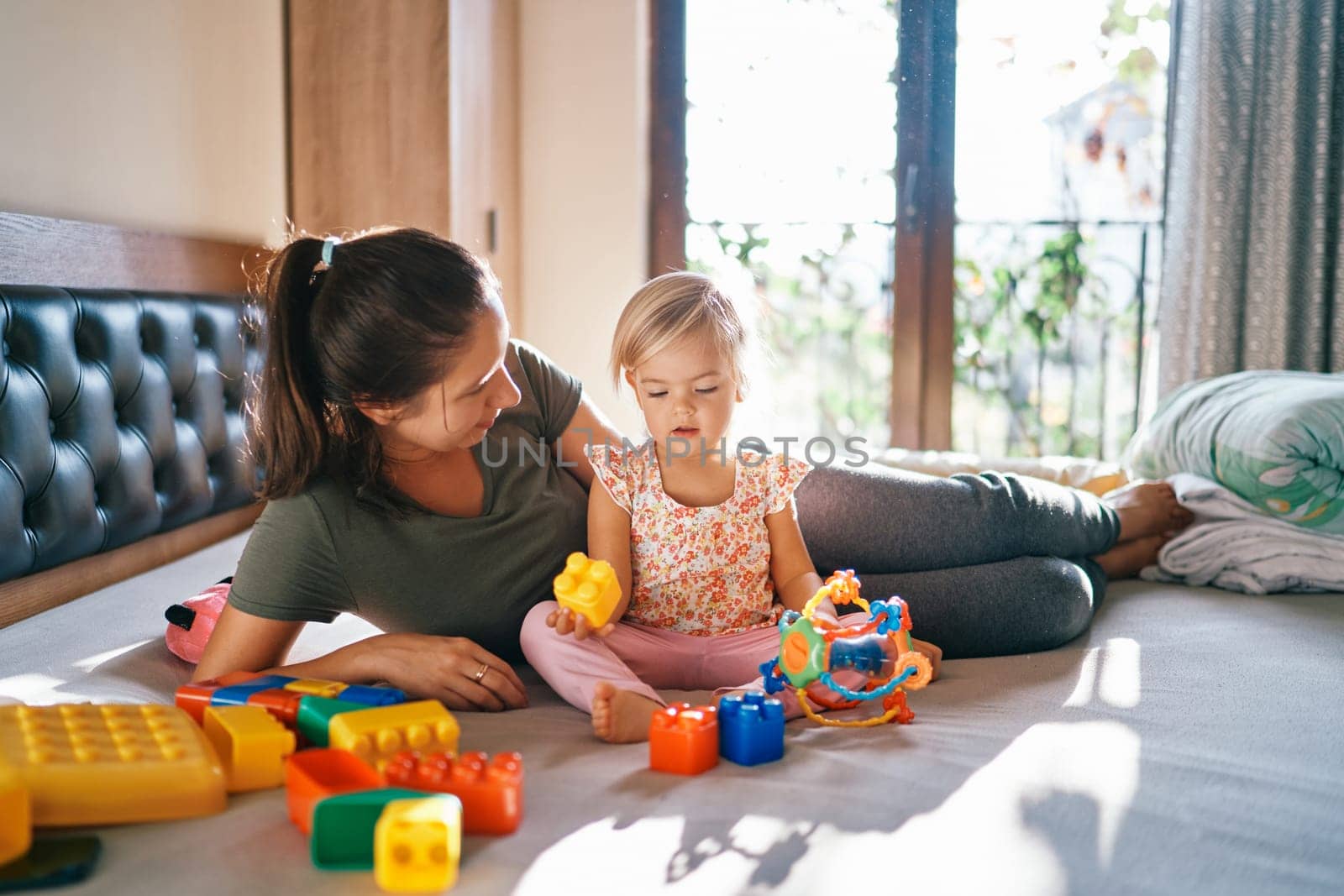 Mom lies next to a little girl on the bed assembling lego by Nadtochiy