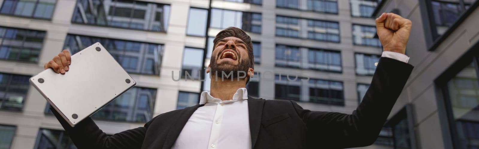 Businessman with laptop standing on office terrace and raised fist is suttisfied of finished project by Yaroslav_astakhov
