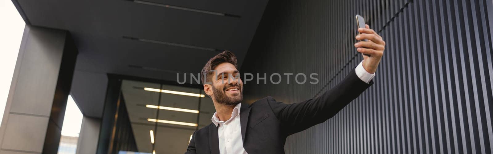 Smiling businessman make a selfie on mobile phone sitting on bench near office building