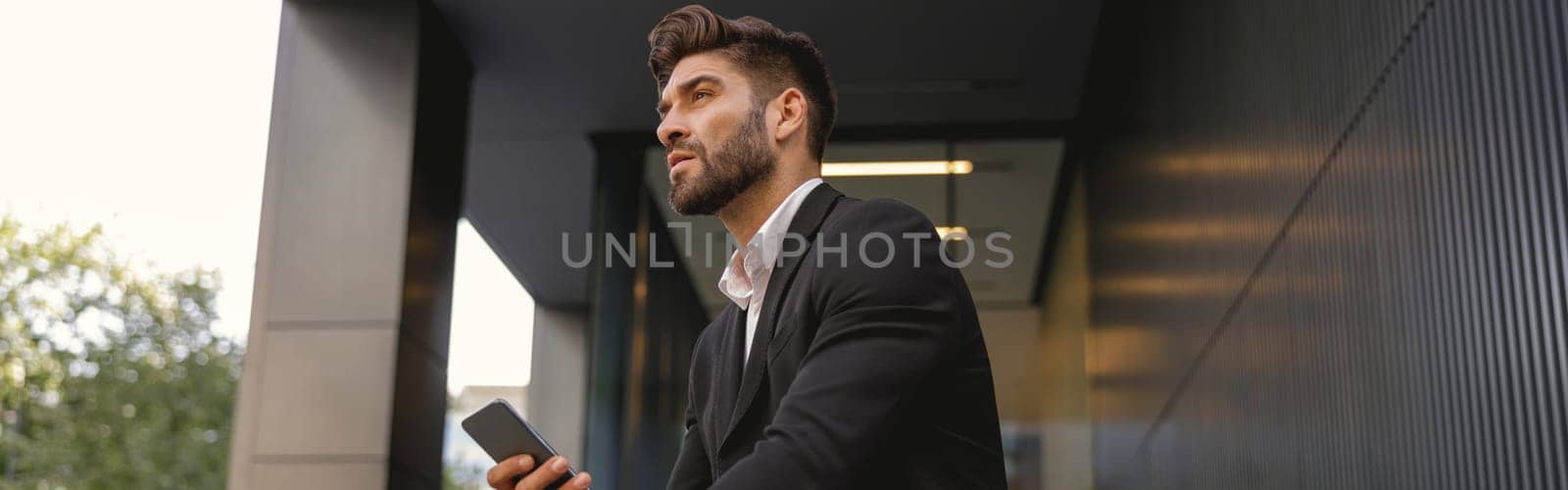 Handsome businessman with mobile phone sitting on bench on office building background and looks away by Yaroslav_astakhov