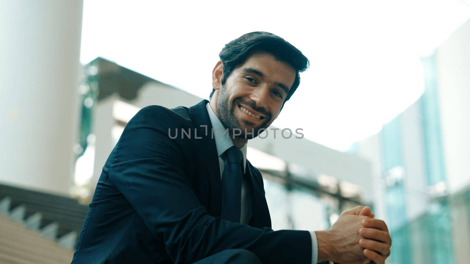 Smiling skilled businessman looking at camera while sitting on stairs. Young professional project manager smile at camera while holding mobile phone at outdoor with blurred background. Exultant.