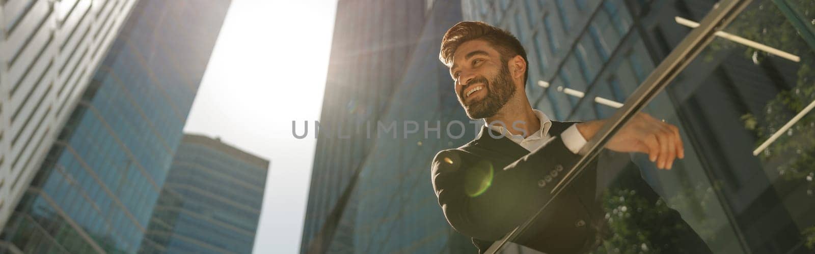 Smiling businessman in suit is standing with laptop on office terrace and looks away by Yaroslav_astakhov