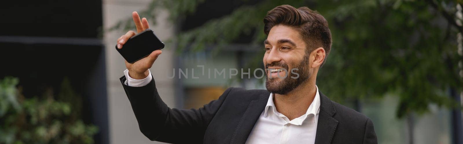 Smiling male manager standing near car with phone and waving hi to colleagues. High quality photo