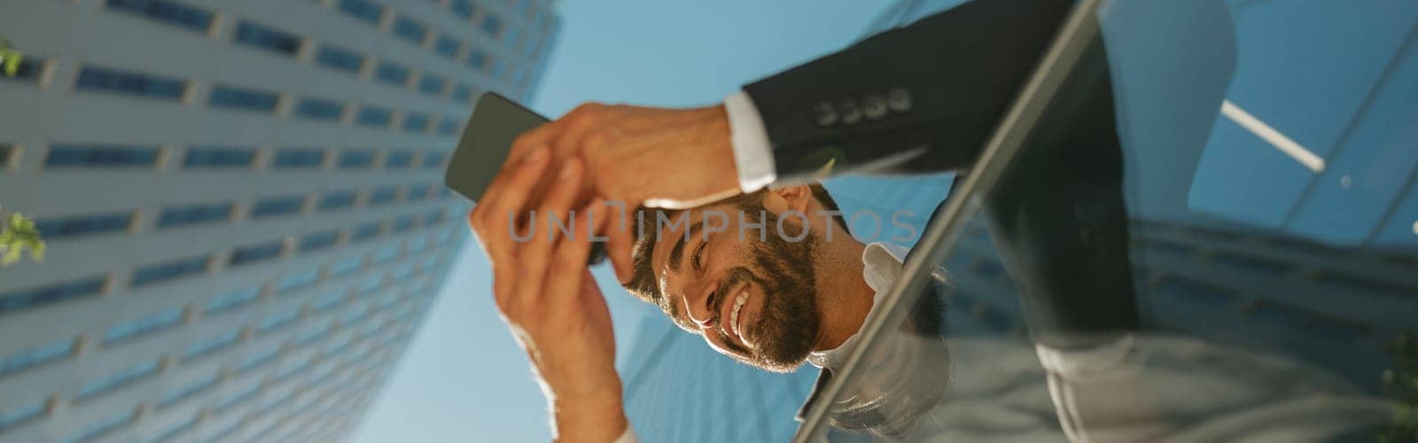Bottom view of handsome entrepreneur using mobile phone standing on background of skyscrapers