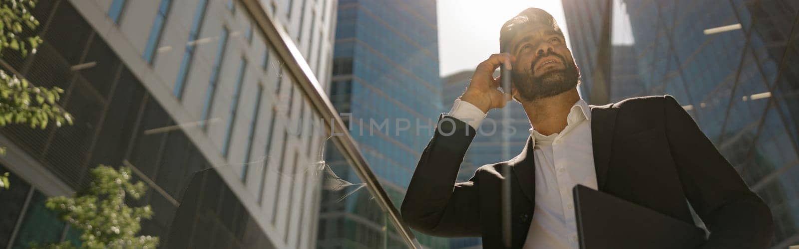 Stylish businessman is standing with laptop on office terrace and looks away. High quality photo
