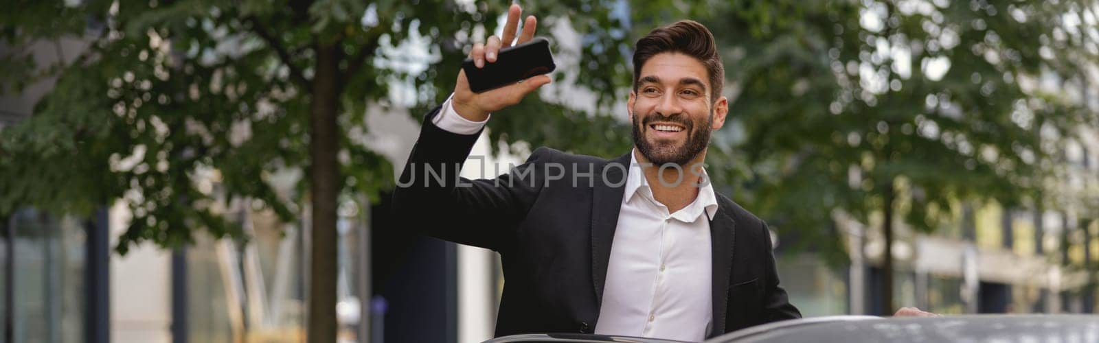 Smiling male manager standing near car with phone and waving hi to colleagues. High quality photo by Yaroslav_astakhov