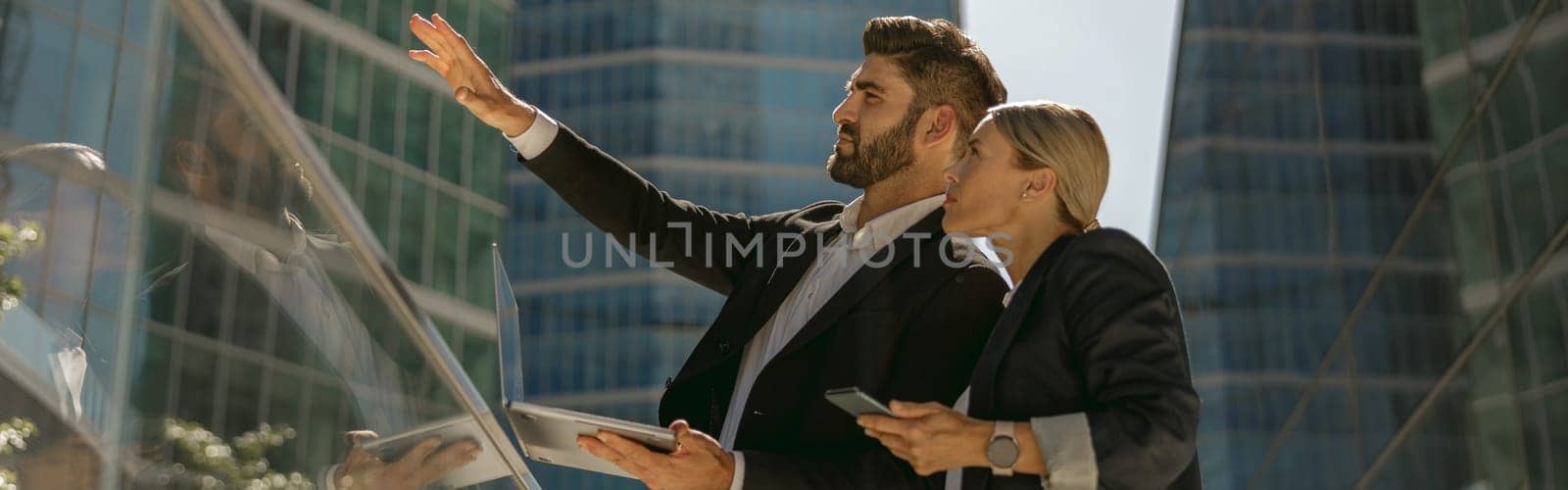 Businessman showing something to his female colleague while standing with laptop on office terrace by Yaroslav_astakhov