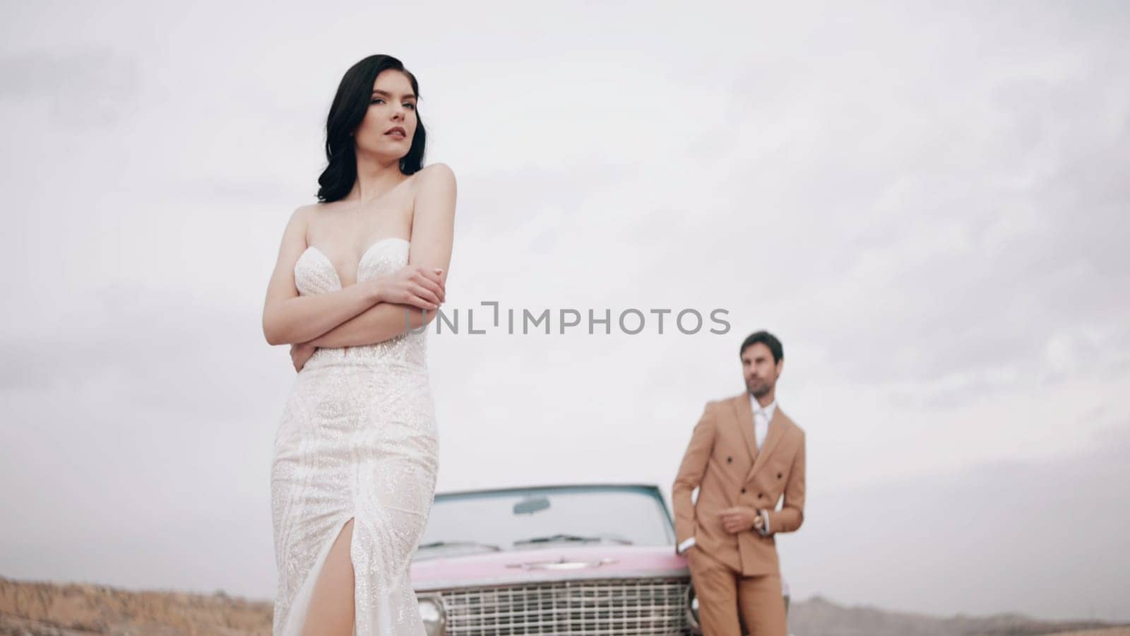 Elegant wedding photo shoot. Action. A beautiful bride in a wedding dress with a groom in a brown suit posing for a photographer on camera with happy faces in the desert next to a pink small car.