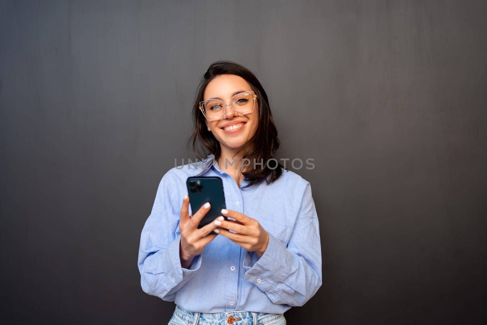 Woman wearing glasses poses with phone against black background. Girl in glasses, dressed blue shirt holding smartphone in hand