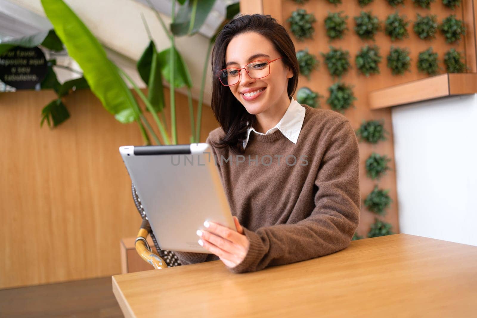 Smiling woman sitting holding digital tablet in hands. Confident woman sits on hall looking at camera holding digital tablet at blur background. Female uses tablet for work