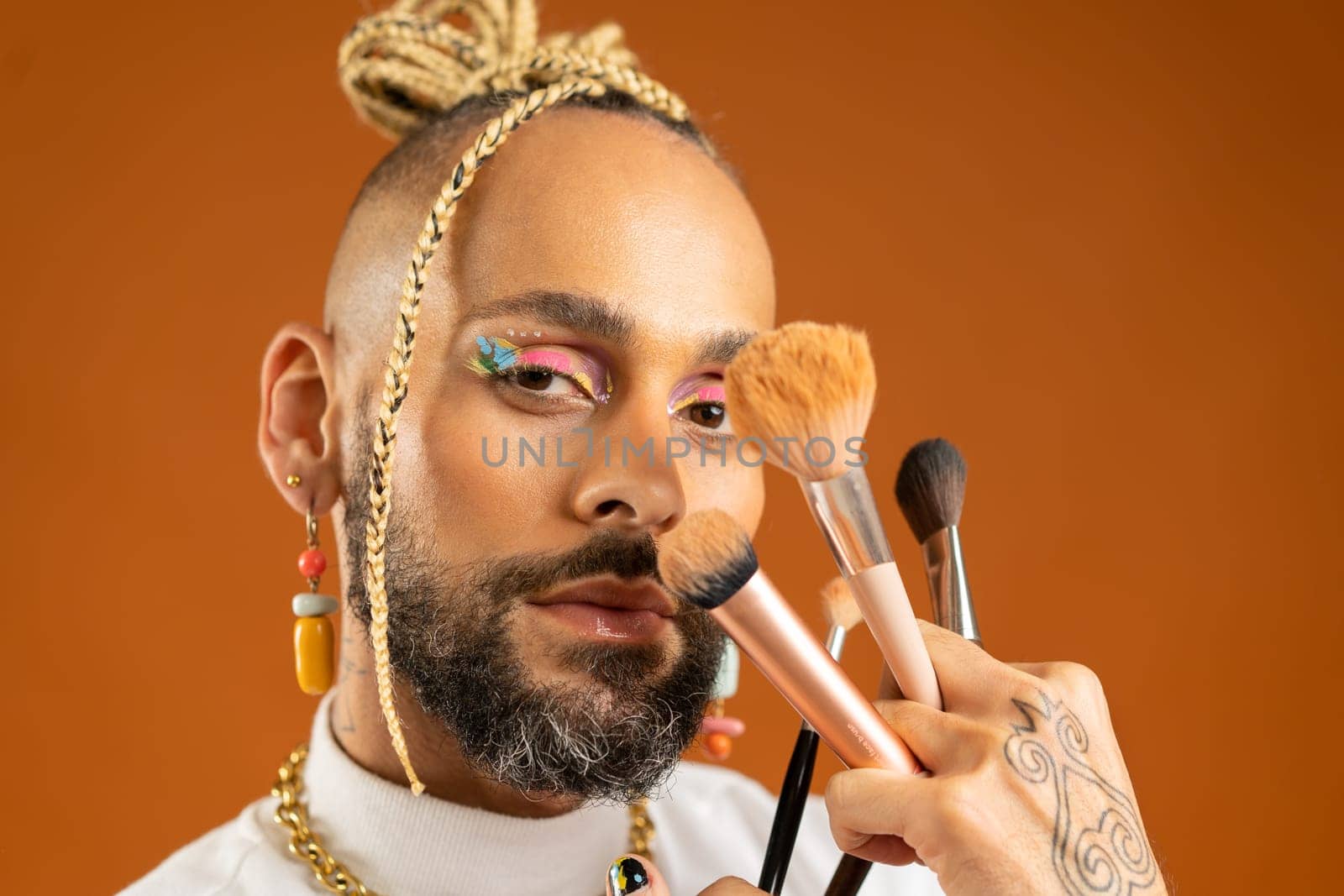 Latino gay make up artist holding makeup brush. Handsome guy wear bright make-up on isolated orange background. Fashion lgbt concept. Handsome makeup artist close up portrait